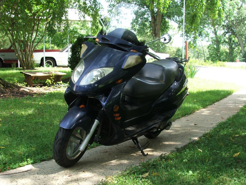Dark blue scooter parked on a sidewalk, front three-quarter view. Green grass and trees surround it
