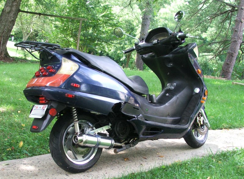 Dark blue scooter parked on a path, rear view showing taillights and exhaust