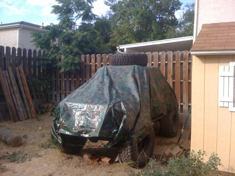 1987 Suzuki Samurai - A vehicle is covered by a camouflage tarp with a spare tire placed on its roof.