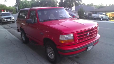 1988 Ford Bronco - This red Ford Bronco, likely a 1992 model based on its grille, is parked curbside. Another SUV is visible in the background.