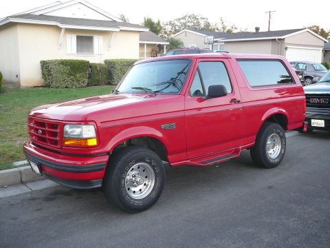 1988 Ford Bronco - This red Ford Bronco, likely from the early to mid-1990s, is parked on a residential street next to another vehicle.