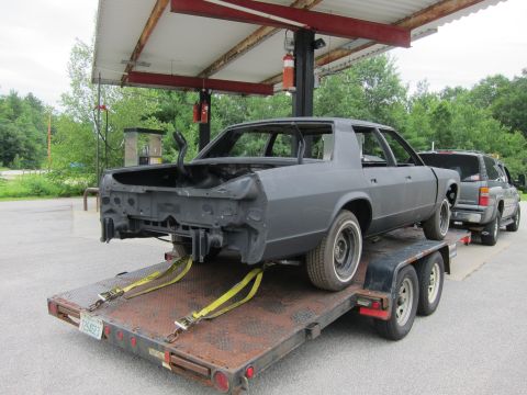 1991 Ford LTD Crown Victoria - A stripped-down car body is loaded onto a trailer, with a pickup truck visible behind it under a covered structure.