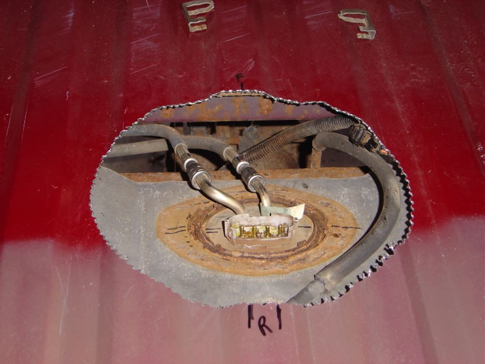 Access hole cut in red floor pan revealing fuel tank sending unit, fuel lines and wiring connector