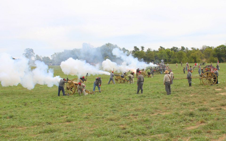 Cannon reenactment with smoke, men in historical attire, and cannons on a grassy field