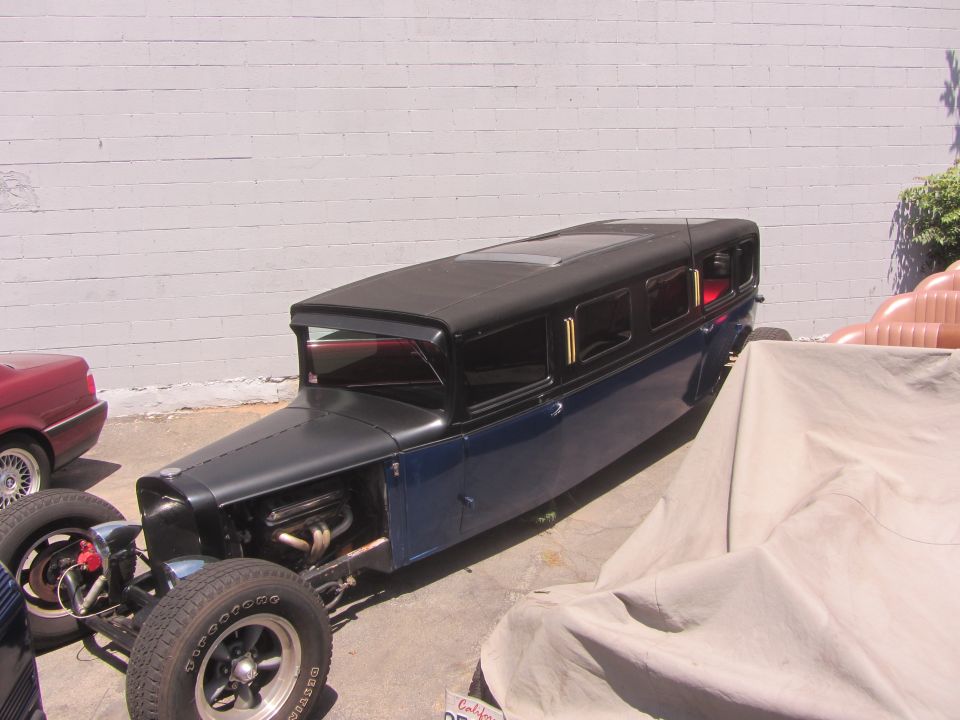 Hot rod limousine with exposed engine, black top, and blue body, parked next to a red car