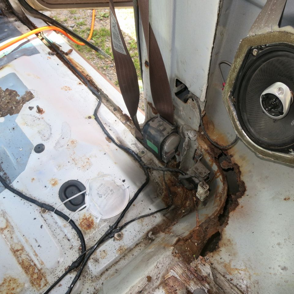 Interior of a vehicle showing severe rust on the floor and seatbelt mechanism. A speaker is visible on the right