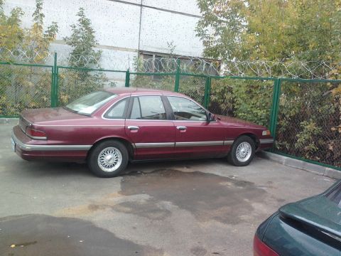 1992 Ford Crown Victoria - Maroon 1992 Ford Crown Victoria parked beside a fence with trees and a building in the background.