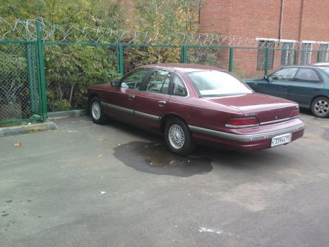 1992 Ford Crown Victoria - A maroon 1992 Ford Crown Victoria is parked in a lot next to a chain-link fence with barbed wire.