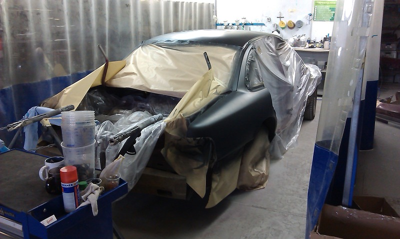 Car body covered in plastic sheeting and drop cloths, undergoing painting in a workshop