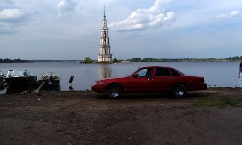 1992 Ford Crown Victoria - A red sedan sits by the water with boats and a tall bell tower in the distance.