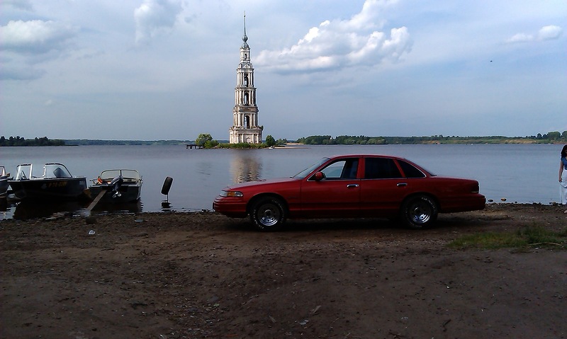 Red sedan parked on dirt by a lake, bell tower in background