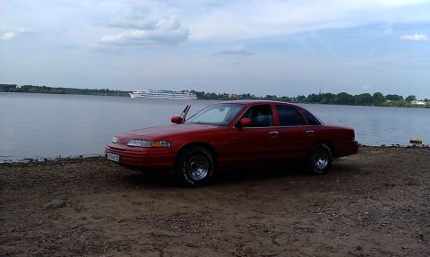 1992 Ford Crown Victoria - A red 1992 Ford Crown Victoria sits parked on a sandy shore next to a wide river with a boat in the distance.