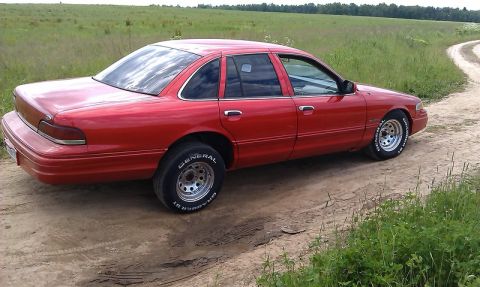 1992 Ford Crown Victoria - A red 1992 Ford Crown Victoria sedan is parked on a dirt road with grassy fields in the background.