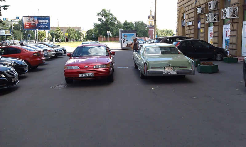 Red Ford sedan parked on street, light green Cadillac sedan parked next to it