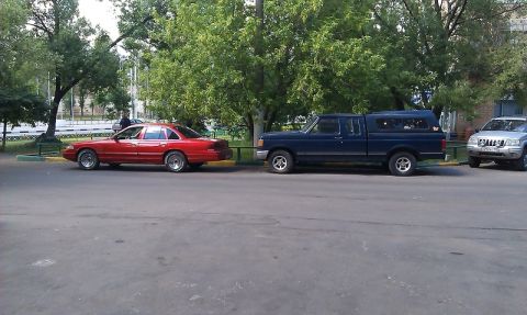 1992 Ford Crown Victoria - A red sedan, a blue pickup truck, and a silver SUV are parked on a street next to trees.