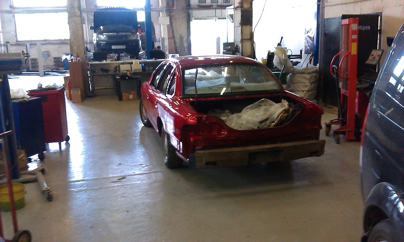 Red sedan with trunk open in a garage. Another vehicle is on a lift in the background