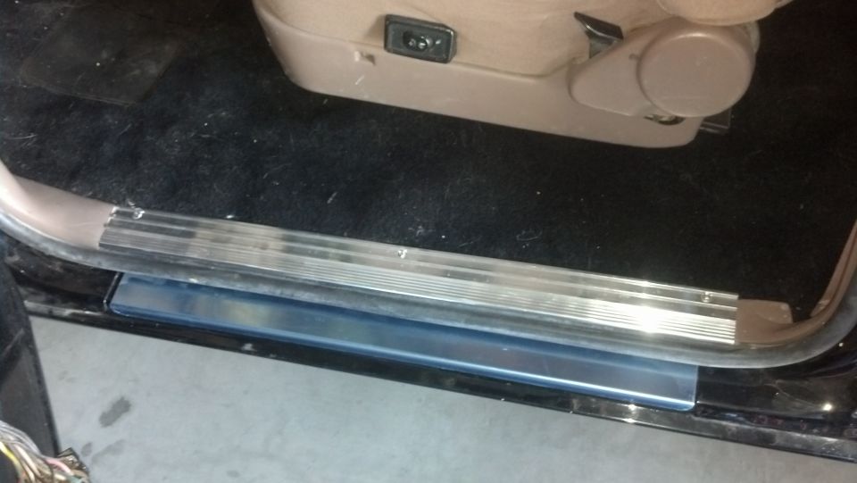 Interior door sill with metal trim and blue plastic protector, showing part of a tan seat with power controls