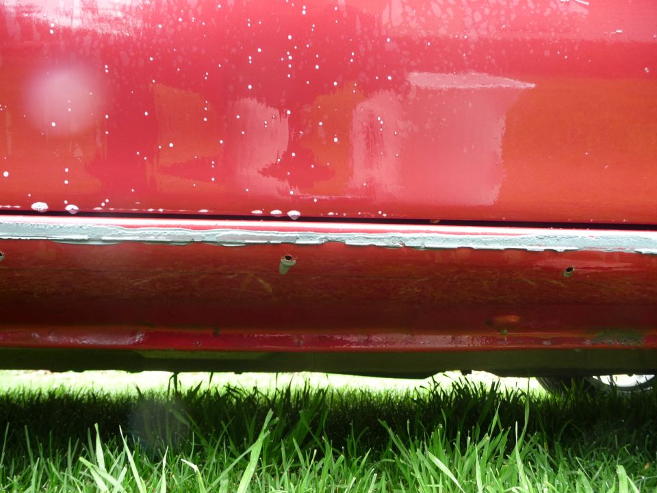 Red car side panel with paint chips and rust along the bottom edge. Some white specks on the paint