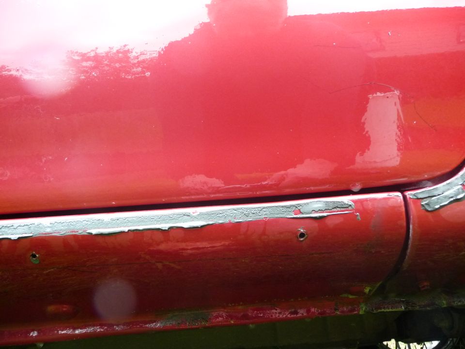 close-up of a red car's lower body panel showing rust and peeling paint