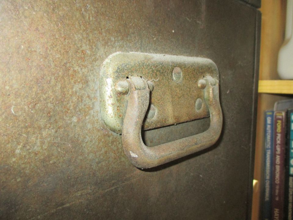 Close-up of a rusted metal cabinet handle on a dark, textured surface. Books are visible on a shelf to the right