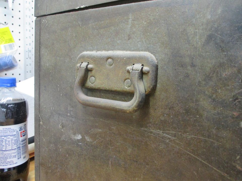 Metal trunk handle on a weathered, scratched metal surface. A bottle of dark liquid with a blue cap is visible to the left