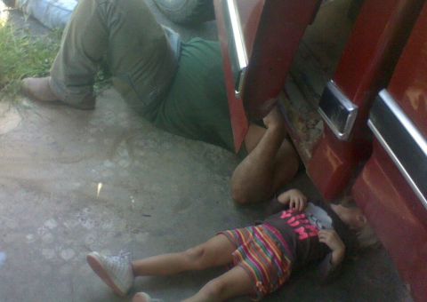 1979 Ford Bronco - A child lies on the ground near a red vehicle, with an adult partially visible working underneath.