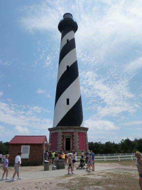 1979 Ford Bronco - A black and white striped lighthouse stands tall under a cloudy sky, with a small red-roofed building and visitors at its base.