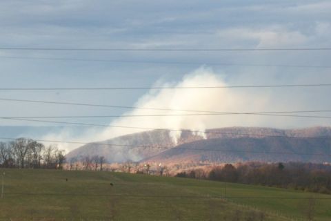 1979 Ford Bronco - Smoke plumes rise from forested hillsides, indicating a fire or controlled burn, under a vast, cloudy sky.