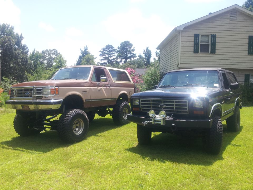 Two lifted Ford Broncos parked on a lawn, one tan and brown, the other black