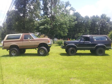 1988 Ford Bronco - Two lifted Ford Broncos, a tan and brown model and a black model, are parked on a grassy lawn.