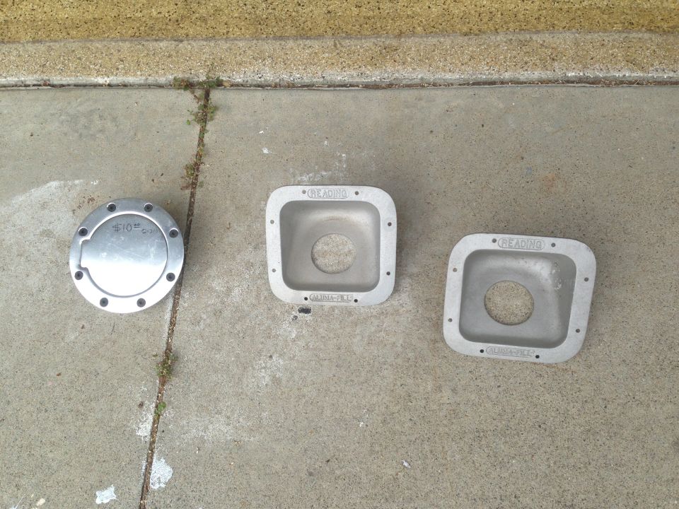 Three aluminum automotive parts, including a circular cap and two square bezels, lie on a concrete surface