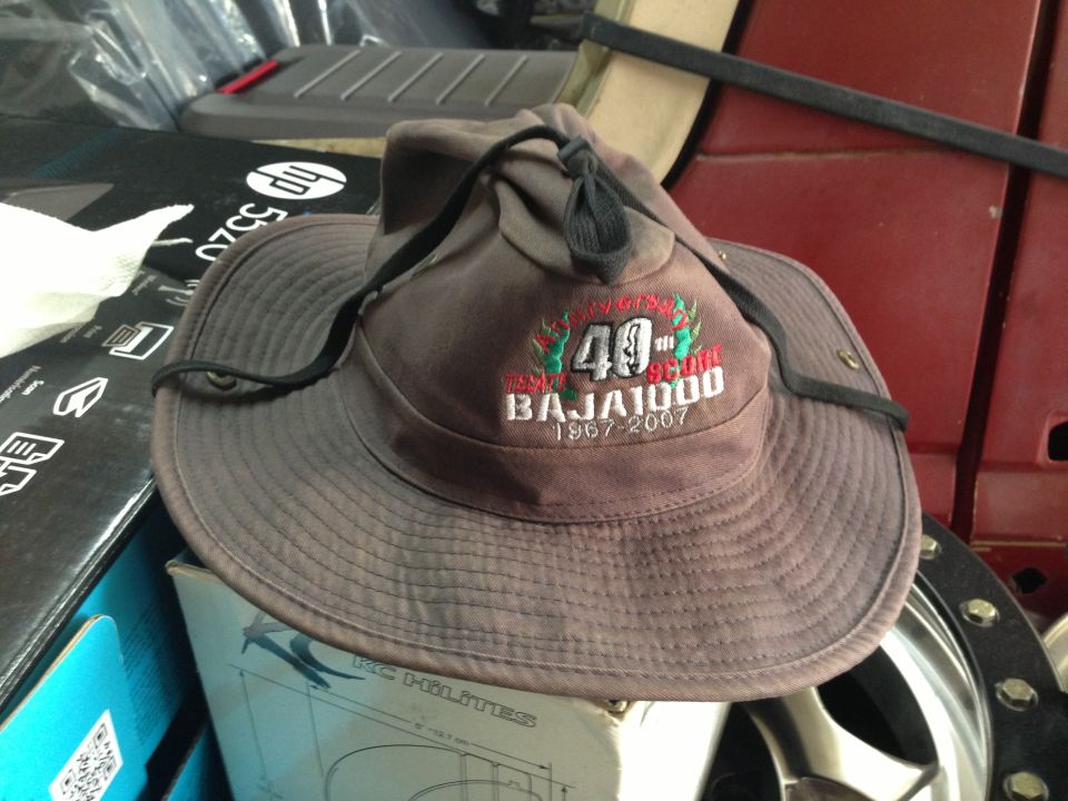 Brown bucket hat with '40th Baja 1000 1967-2007' embroidery sits on boxes next to a red vehicle interior