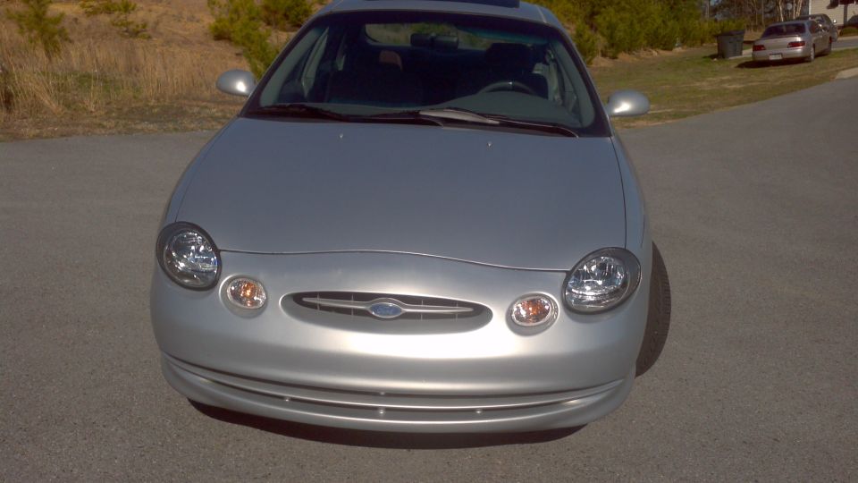 Front view of a silver Ford sedan, possibly a Taurus from the early 2000s. Another car is parked on the side of the road in the background