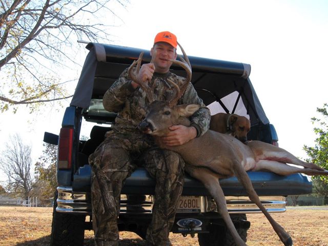 Man in camo with harvested buck on tailgate of blue Ford Bronco SUV, dog in back