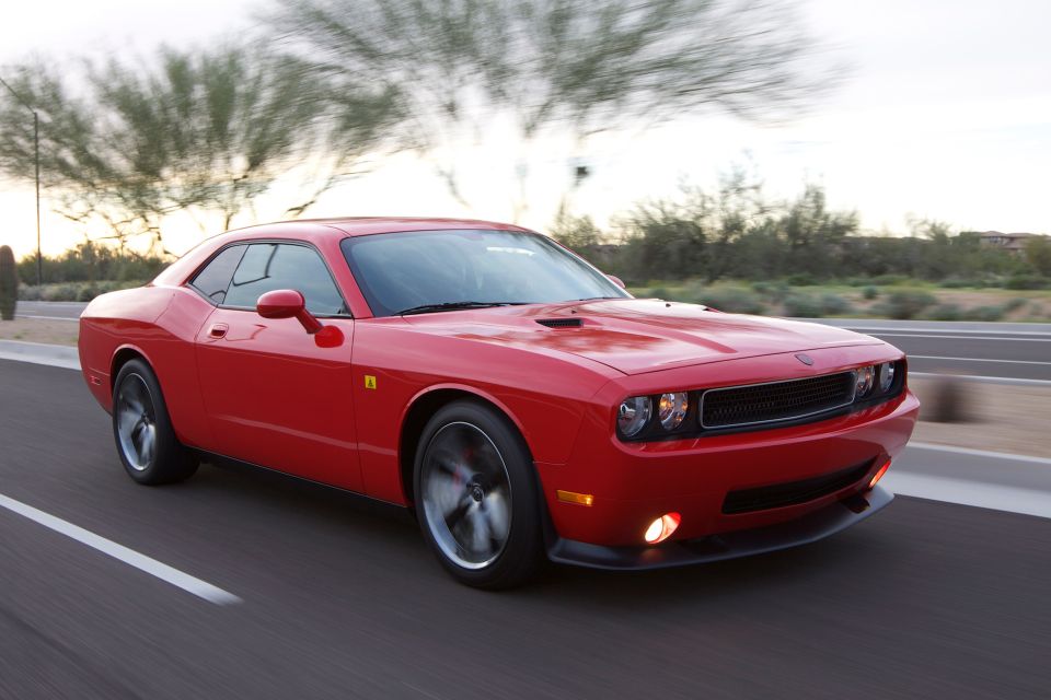 Red Dodge Challenger driving on a road, front 3/4 view. Hood scoops visible
