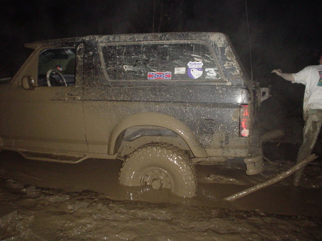 Mud-covered Ford Bronco stuck in deep mud at night, person pulling tow strap