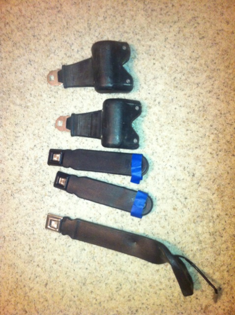 1982 Ford Bronco - A set of black seat belts, some with blue tape on the buckles, are laid out on a speckled surface.