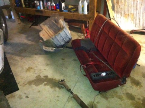 1982 Ford Bronco - A red velour bench seat and a large metal component are on the floor of a garage, with shelves of supplies in the background.