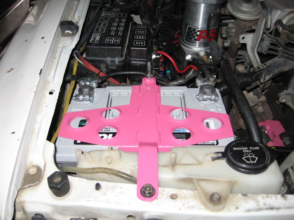 Pink airplane-shaped battery mount over a car battery in an engine bay