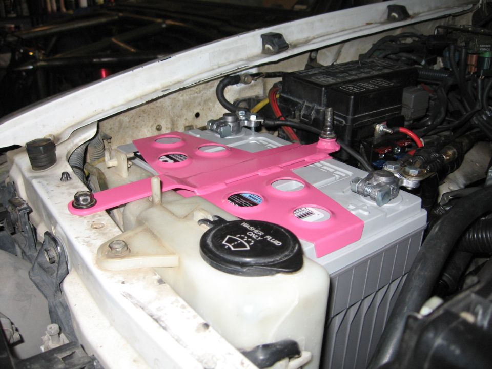 Pink battery hold-down bracket over a gray battery, with a white washer fluid reservoir below