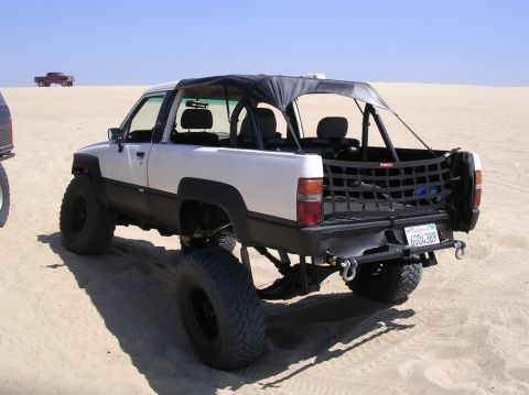 1983 Ferrari Enzo - Modified pickup truck with a soft top, seen on a sandy terrain. Another vehicle is in the background.