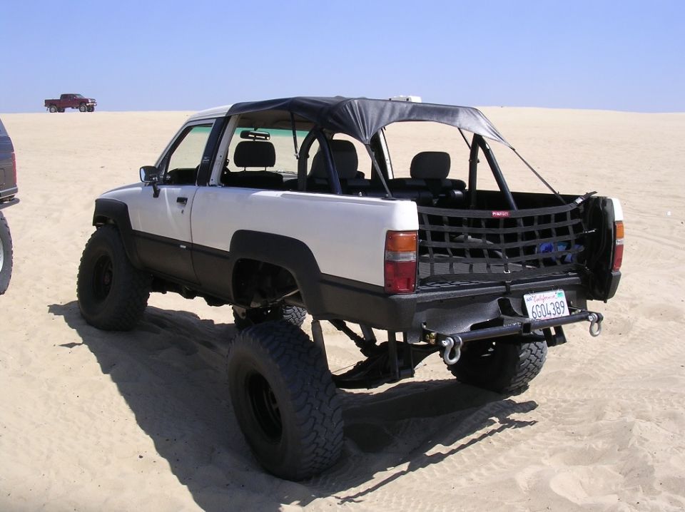 Lifted white and black pickup truck with a soft top, modified for off-road use, parked on sand. Another red pickup is visible in the distance