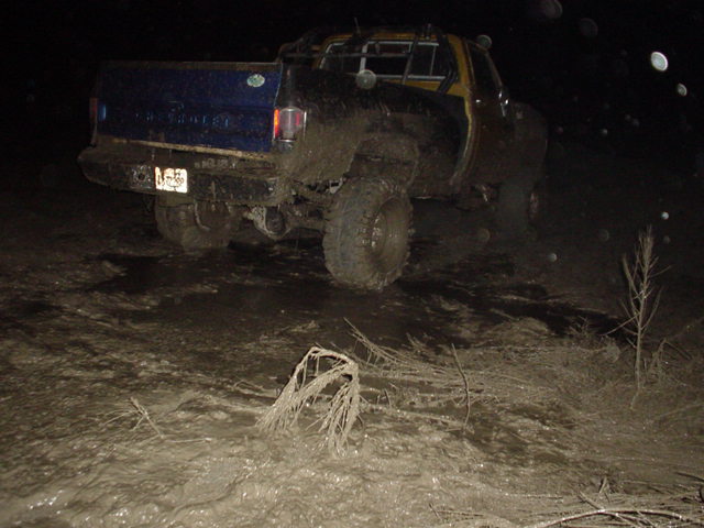 Blue lifted SUV covered in mud at night in a muddy field, rear three-quarter view