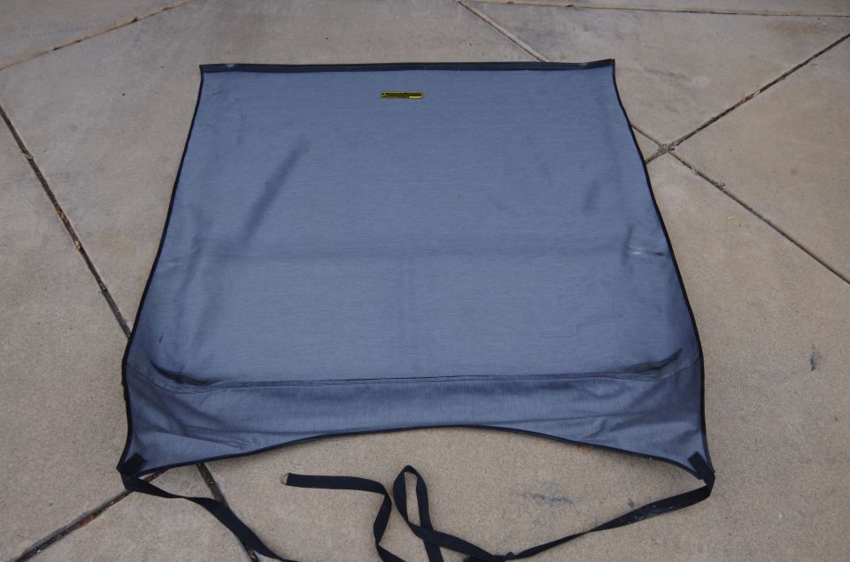 Blue fabric convertible top with black trim and straps, laid out on a concrete surface. A yellow tag is visible