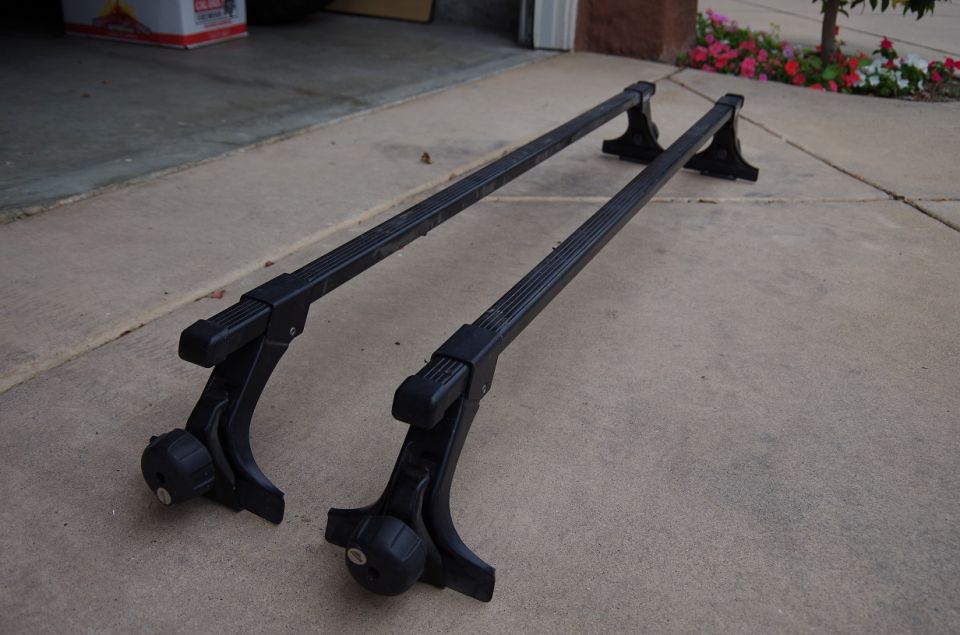 Pair of black roof rack crossbars with adjustable mounts, resting on a concrete driveway