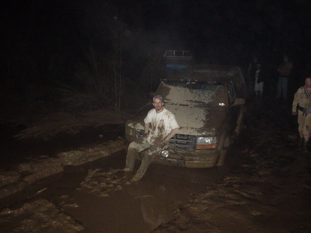 Mud-caked Ford 4x4 stuck in a deep mud hole at night with a person sitting on the bumper