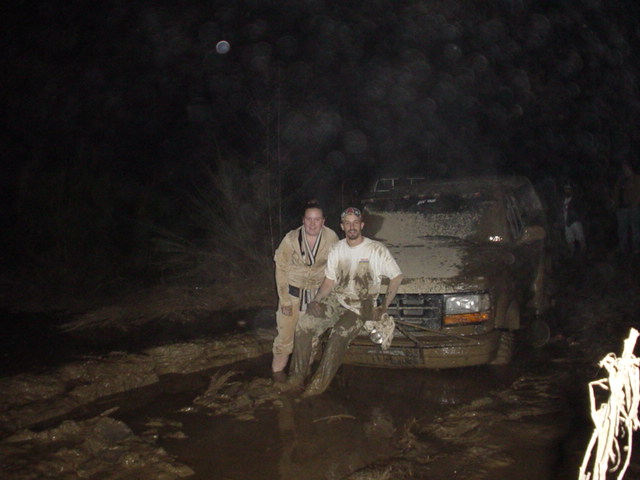 Two men standing in mud beside an SUV stuck in a muddy trail at night