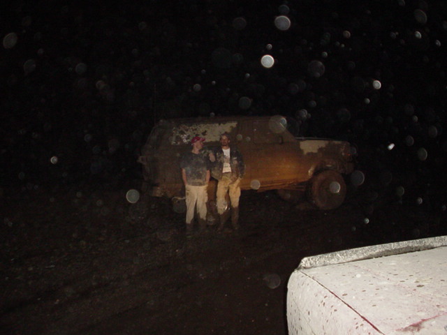 Two muddy people at night standing beside a heavily mud-caked full-size SUV