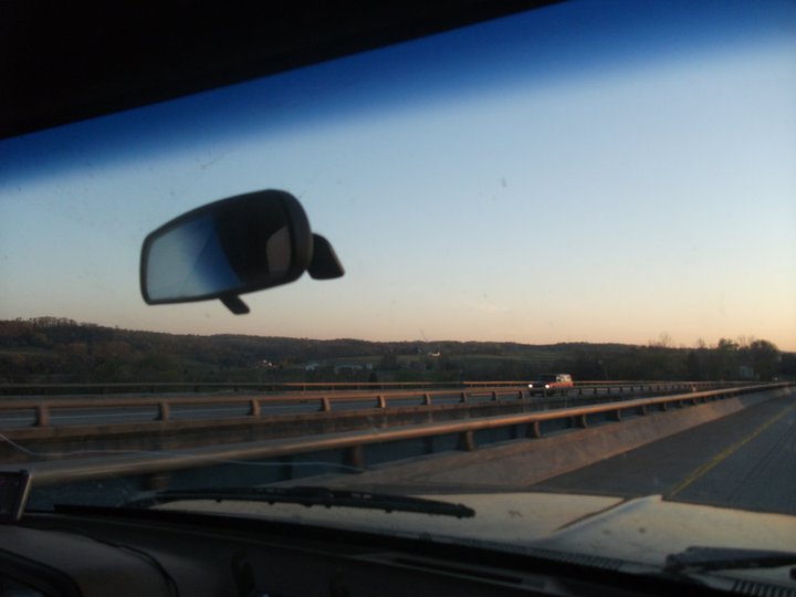 View from inside a vehicle looking out the windshield at a highway overpass and distant hills at sunset. A side mirror is visible