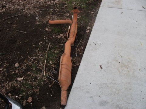 1990 Ford Bronco - A rusty exhaust system part lies on the ground next to a concrete walkway.